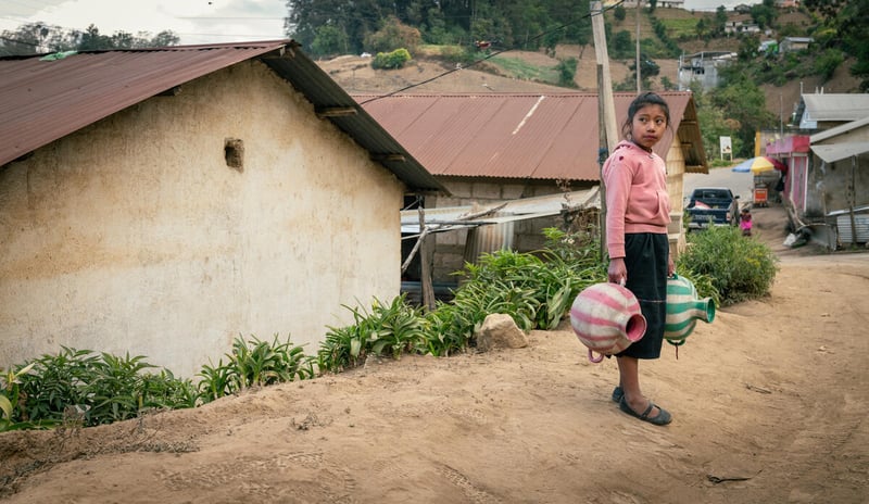 A girl holds a clay water jug while walking down a dirt road.
