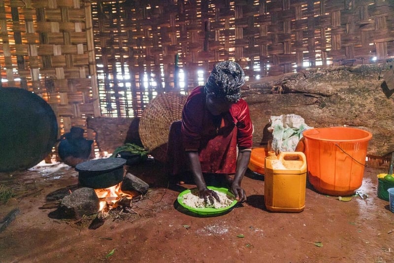 A woman kneels on the floor in her house over a plastic basin as she uses her hands to mix water into grains. There is a pot on a fire next to her and a bucket and jerrycan on her other side.