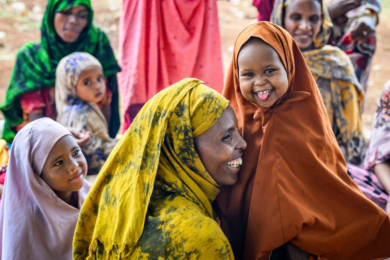 A girl smiles and sticks her tongue out while a woman embraces her. Other girls and women look on.