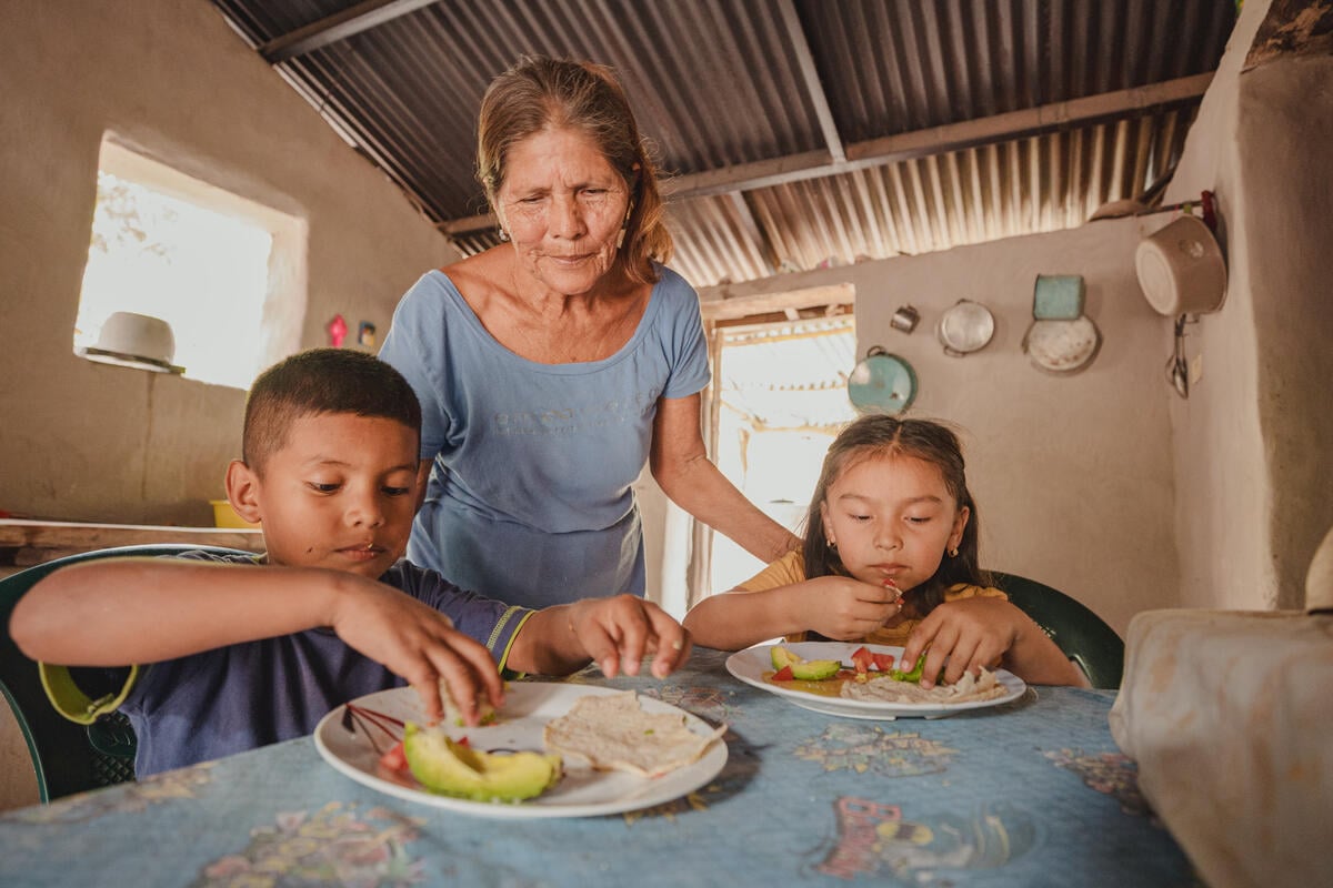 A woman stands between a boy and a girl seated at a table, watching them as they eat. Sunlight streams into the adobe-style home.