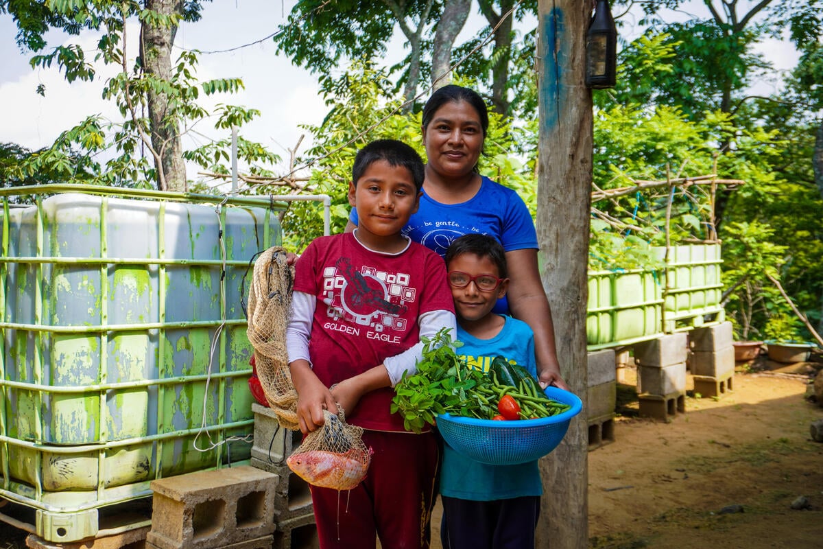 A woman and two children stand in front of large plastic bins used to grow vegetables under trees.