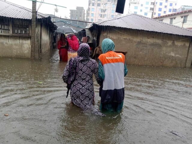 Two women, one an aid worker, wade arm-in-arm through floodwater toward an opening between two rows of single-story concrete homes. Other women walk ahead holding umbrellas.