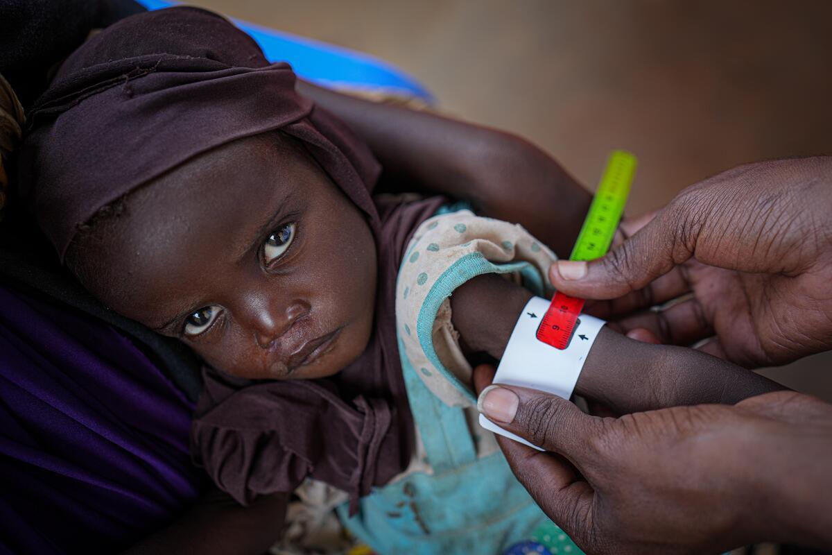A health worker measures a child’s mid-upper arm circumference (MUAC) to screen for malnutrition. The measurement falls in the red zone, indicating severe acute malnutrition.