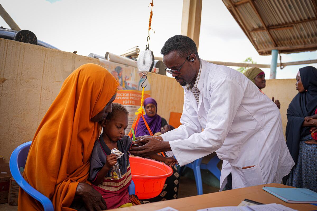 A healthcare worker measures a child’s upper arm to screen for malnutrition. The child sits on their mother’s lap at an open-air health care clinic.