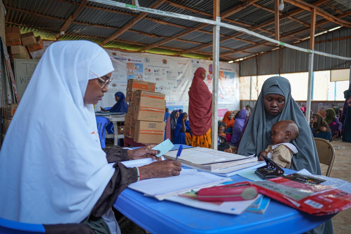 Women in long head coverings seated at a table, one holding a young child and the other writing in a notebook in a clinic.