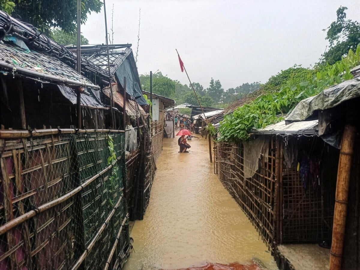 Floodwaters rise to the doorsteps of bamboo houses with their roofs covered in lush vegetation. A person in the distance holds an umbrella. A small crowd gathers at the far end of the row.