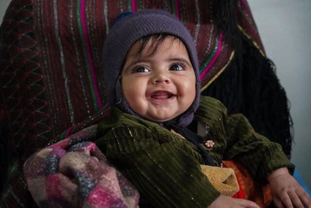 A smiling four-month-old girl sits on her mother’s lap, bundled up in a cold-weather jacket, cap, and blanket.