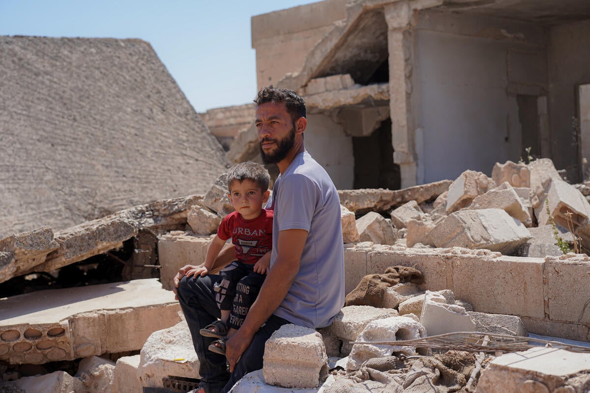 A bearded man holds his young son on his lap amid the rubble of their destroyed home with broken concrete blocks and slabs piled behind them.