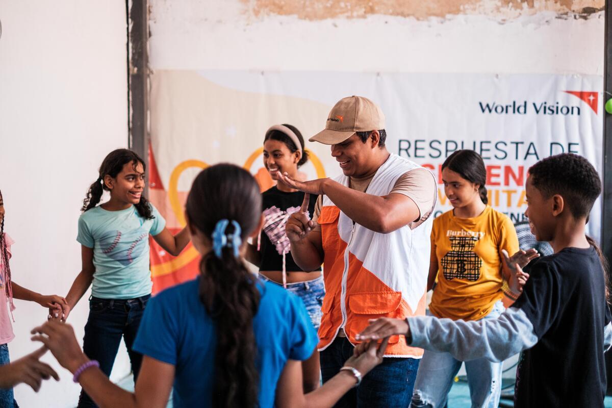 A man wearing an aid worker vest smiles while making hand gestures in the middle of children holding hands and playing. A banner hangs behind them.