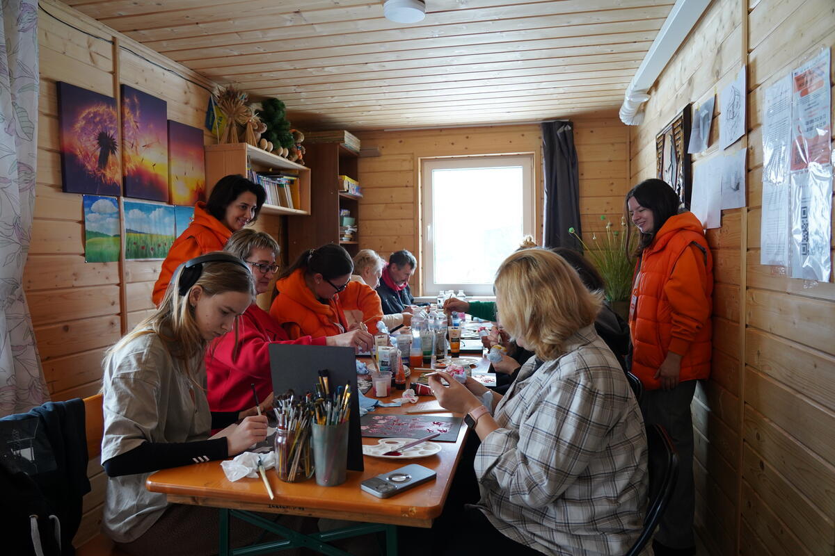 Aid workers stand behind people around a table doing crafts inside a narrow room with wood panels adorned with pictures and paintings.