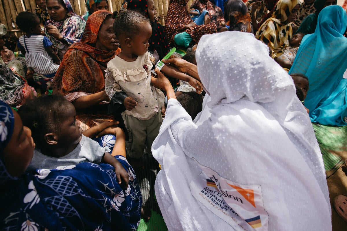A healthcare worker measures a child’s Mid-Upper Arm Circumference (MUAC) as women and children gather around them. The measurement shows green, meaning an acceptable nutritional status.