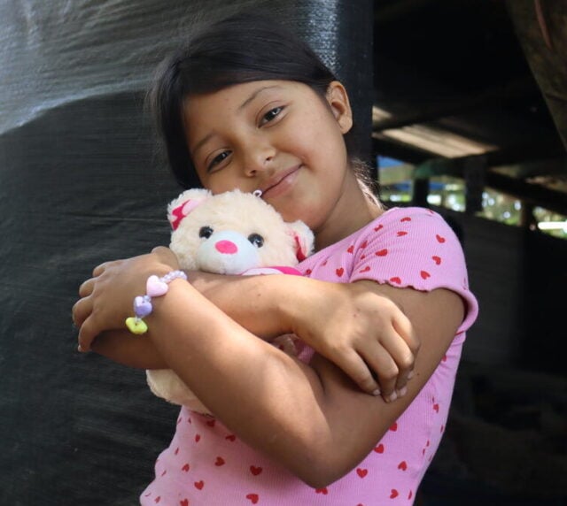 A girl in a T-shirt printed with hearts wears a bracelet of hearts and hugs a teddy bear.