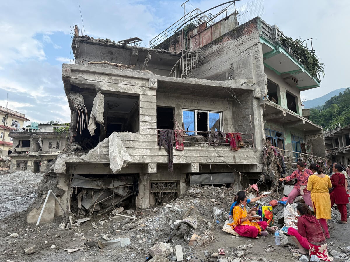 Women sit outside a damaged multi-story building with exposed concrete and rebar following catastrophic flooding.