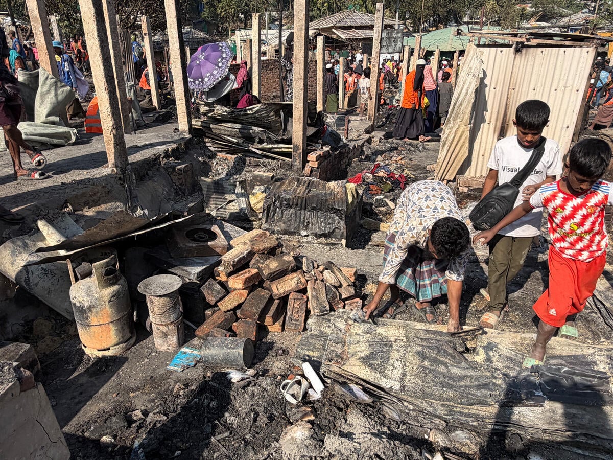 People step over charred remains of a dwelling. A crowd is seen in the distance between wooden pilings.