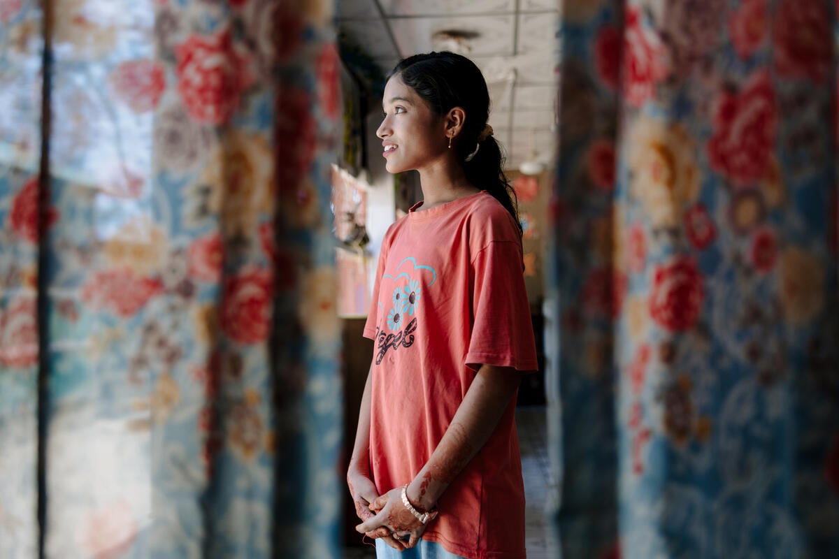 Profile of a girl with a long ponytail standing near an open window, framed by floral curtains.