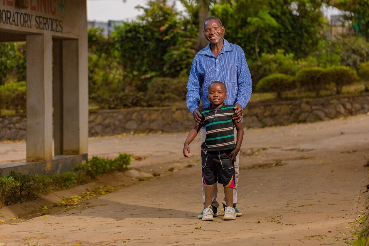 A man stands behind a boy with his hand on the boy's shoulders. Both look at the camera and smile. The boy has prosthetic legs.