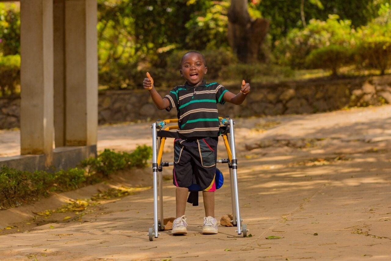 A boy stands inside a metal walker. He holds his hands up with both thumbs up, mouth open as he shouts for joy.
