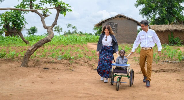 A man and a woman each use one hand to push a child in his wheelchair through the dirt. There are two thatch-roof buildings behind them and a tree to their right.
