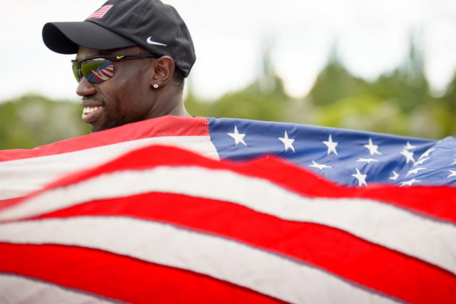 A man smiles and wears an American flag around his shoulders.