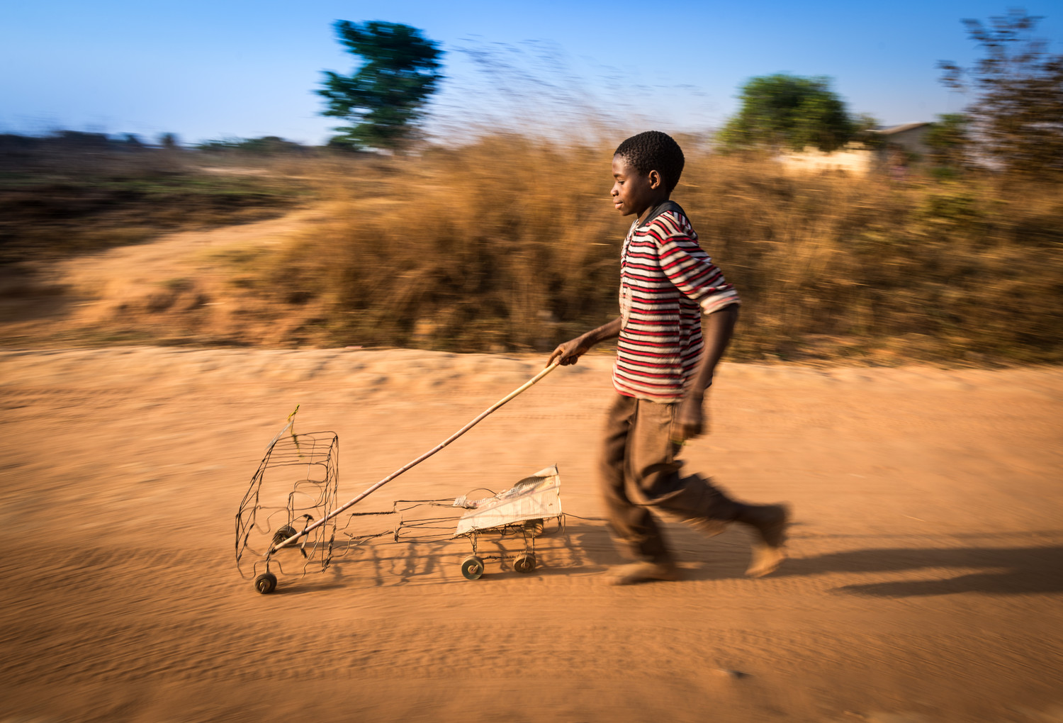 Felix Kansapato, 13, pushes a toy truck made out of wire and cardboard down a road in Kapululwe, Zambia. Felix created something from nothing, using only the materials available: scrap pieces of wire, cardboard, and old wooden wheels. ©2015 Eugene Lee/World Vision